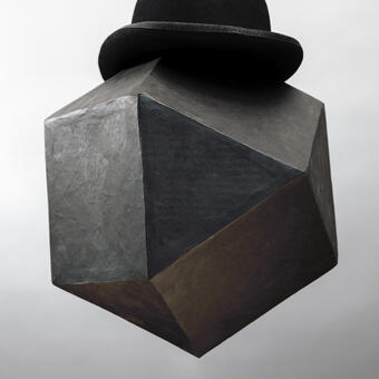 Abstract portrait of a black geometric form made from papermache wearing a black derby cap. Photographed in studio against a gray background.