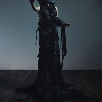 Portrait of a very tall pale-skinned woman with a large black dodecahedron shaped head holding three fingers up on her right hand. This was shot with practical effects in studio and color graded in post. I wanted it to feel otherworldly like a dream, but remain anchored to our actual real life production space. If you look closely you will see gaffer's tape on the floor, marking where we typically run autopoles for seamless backgrounds, and nail holes / wear and tear in the drywall backdrop. 