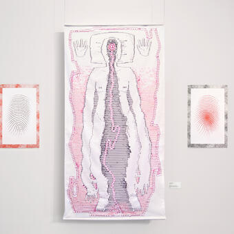 A triptych featuring a floating, white, cotton tapestry in the center, hanging by clear wire 2 inches from the wall, with two smaller risograph prints pinned on either side. Composed of printed black and magenta computer symbols with overlaid embroidery and beadwork, a humanoid figure laying on a pillow features a large gash resembling a cave or portal that opens from their forehead down to their limbs, which appear glued together. Inside the gash is a large “at” symbol, from which a magenta cord reminiscen