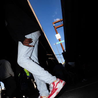 A man's legs lit in the foreground, crossed during a breakdancing session, a triangular sliver of light in the background revealing a sign that says "Inner Harbor"