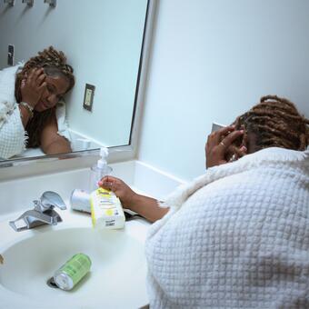 This photo shows a young woman sitting on a bathroom floor wrapped in a white blanket, leaning against the wall and sink while holding a bag of sugar. The scattered items around the sink and her tired posture suggest stress, exhaustion, and the physical effects of sugar withdrawal. Her reflection in the mirror adds to the feeling of vulnerability and struggle.