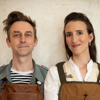 Two people wearing leather and canvas aprons, photographed against a textured light wall. On the left, a man with tousled brown hair in a striped shirt and denim vest with a small enamel pin. On the right, a woman with dark hair pulled back, wearing a white striped blouse with a metal brooch and drop earrings.