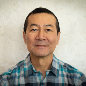 Man with short dark hair wearing a teal and gray plaid shirt, photographed against a textured light wall.
