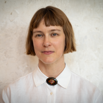 Woman with a short brown bob and bangs, wearing a white collared shirt with a decorative oval brooch, photographed against a textured light wall.
