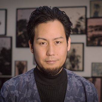 Man with dark swept-back hair and a goatee, wearing a black turtleneck and a blue floral-patterned jacket, photographed in front of a wall hung with framed photographs.