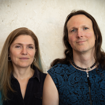 Two people photographed together against a textured light wall. On the left, a woman with long blonde hair wearing a black sleeveless top and hoop earrings. On the right, a man with long dark hair wearing a blue patterned top and a black bead necklace.