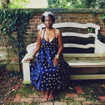 An image of Pamela Woolford in a long, formal, blue dress, barefoot and seated on a large white bench in front of a brick wall with greenery around. 