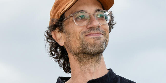 Bruce Willen, looking slightly up and to the right, wearing glasses, and orange baseball hat and black shirt, against a background of sky