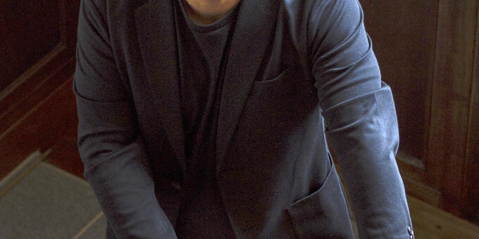 Lysley Tenorio, clad in hat, glasses, and navy blazer, standing in stairwell, looking directly at viewer