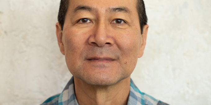 Man with short dark hair wearing a teal and gray plaid shirt, photographed against a textured light wall.