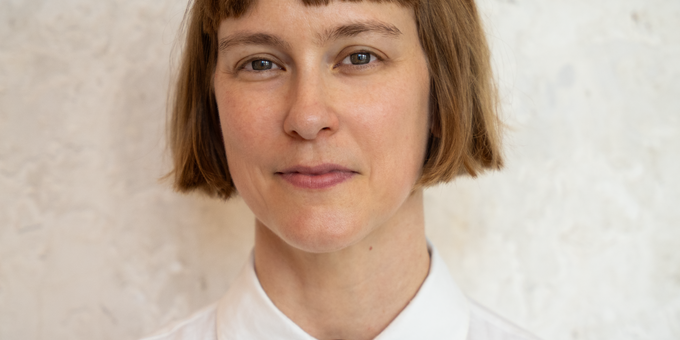 Woman with a short brown bob and bangs, wearing a white collared shirt with a decorative oval brooch, photographed against a textured light wall.