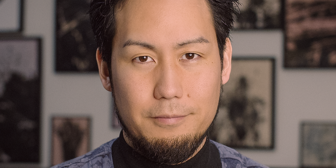 Man with dark swept-back hair and a goatee, wearing a black turtleneck and a blue floral-patterned jacket, photographed in front of a wall hung with framed photographs.