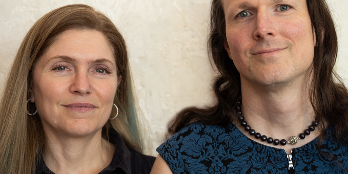 Two people photographed together against a textured light wall. On the left, a woman with long blonde hair wearing a black sleeveless top and hoop earrings. On the right, a man with long dark hair wearing a blue patterned top and a black bead necklace.