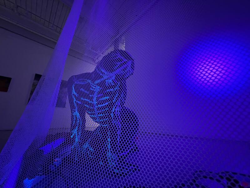 a person wearing a full body suit covered in a glowing nervous system outline crawl an the floor behind a mesh scrim.