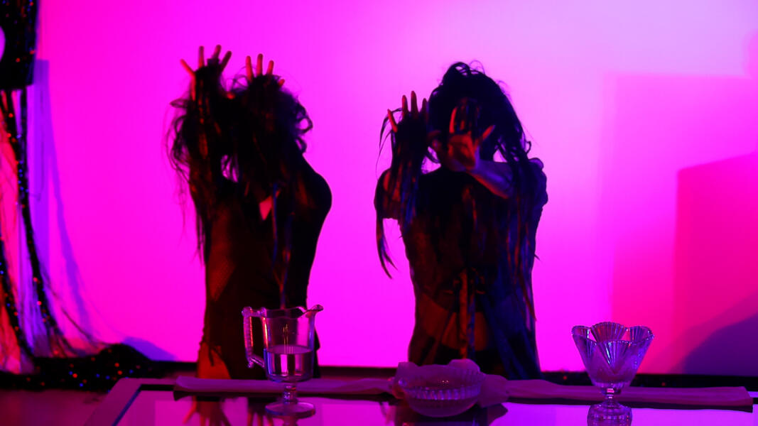 Red and Rain performing an ancestor ritual in the gallery installation reaching fingers through long black wigs worn over their faces. 