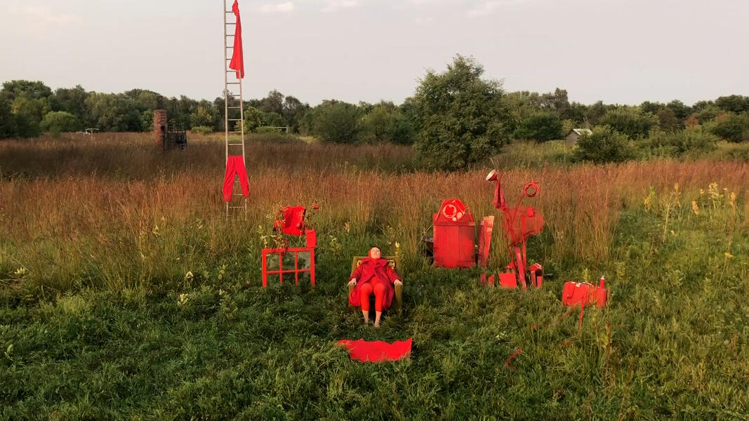 Red is wearing all red and seated in a chair in lush field with red objects surrounding them in the grass and hanging on a tall ladder behind them. 