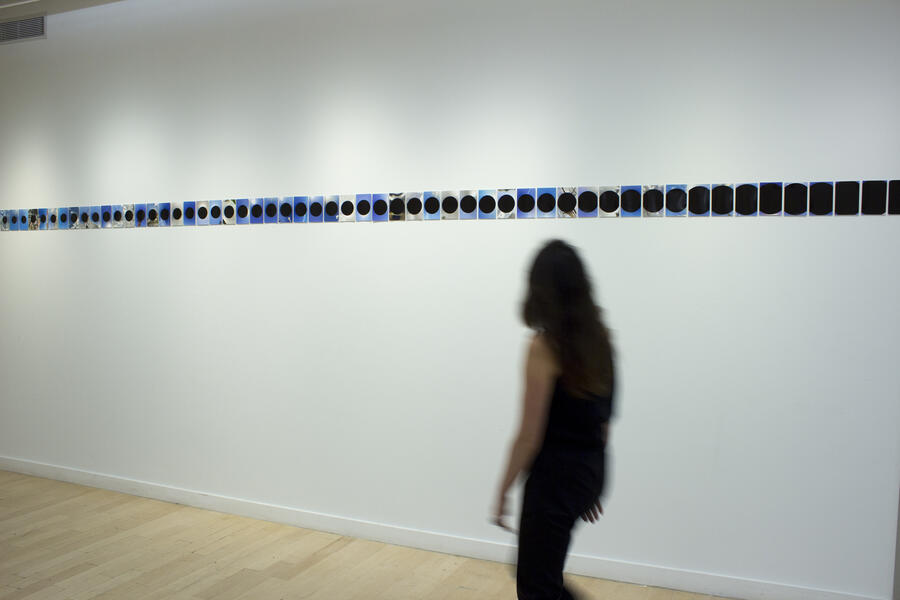 Long row of small images on a white wall with slightly blurred person walking past