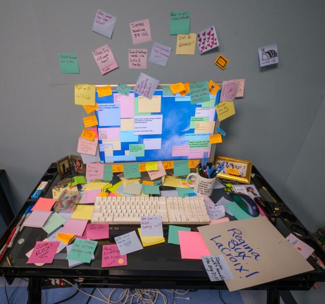 A computer screen and keyboard sit on a glass desktop, with virtual sticky notes covering the screen and physical sticky notes covering the computer and desktop area