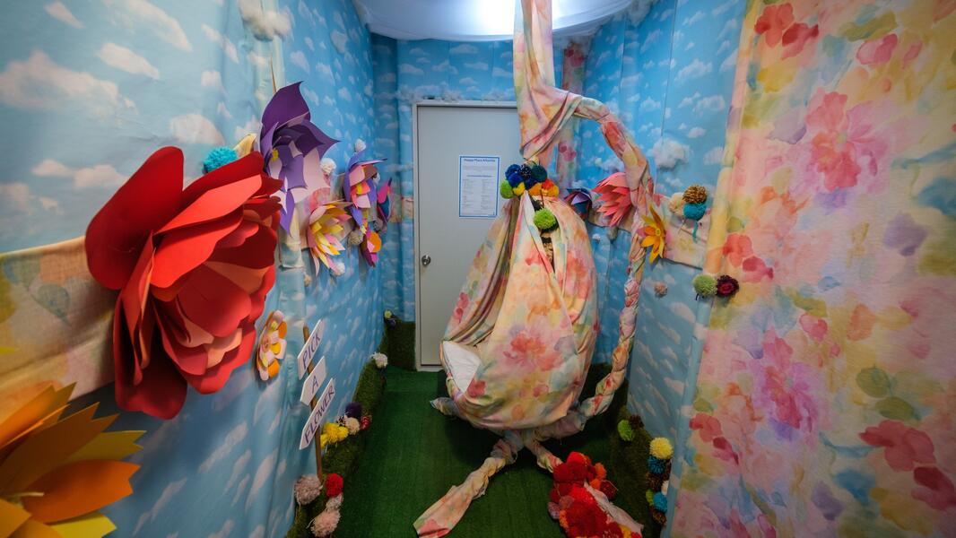 A small room with a swing chair covered in floral fabric, with wallpaper covered in clouds and colorful paper flowers adorning the walls