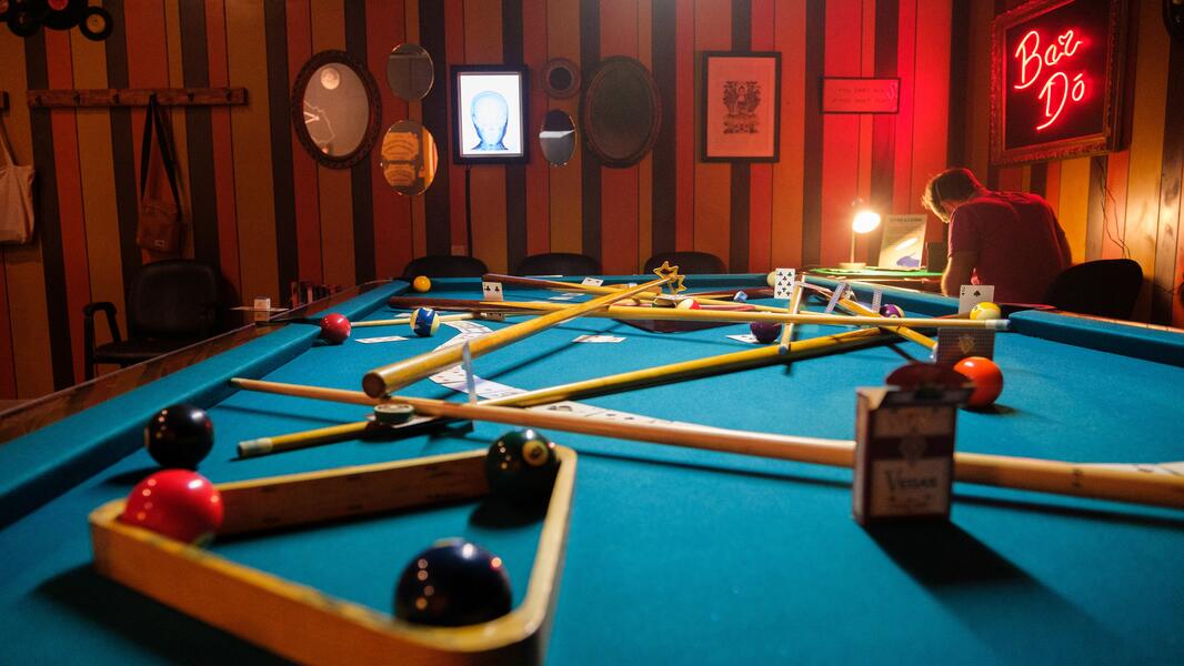 The top of a billiard table, covered in billiard balls, pool cues and playing cards. The wall of a dimly lit bar, covered in mirrors and framed artwork, is in the background.