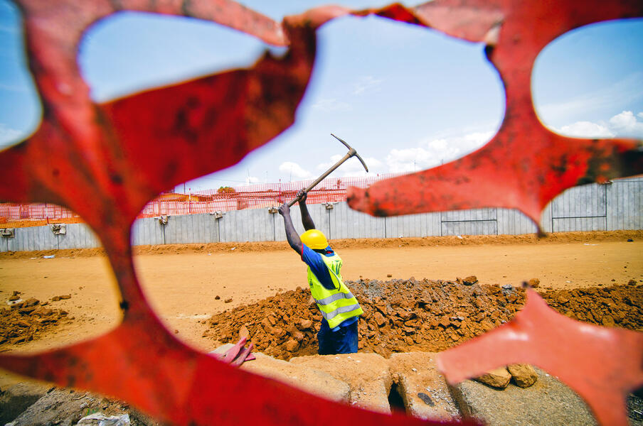 Captured through the visual crest of an orange guardian safety fence at the peek of the action with a sense of determination, a man labors to dig the foundation of one the pillars of the 3rd major bridge in Abidjan, Ivory Coast, connecting the North and t