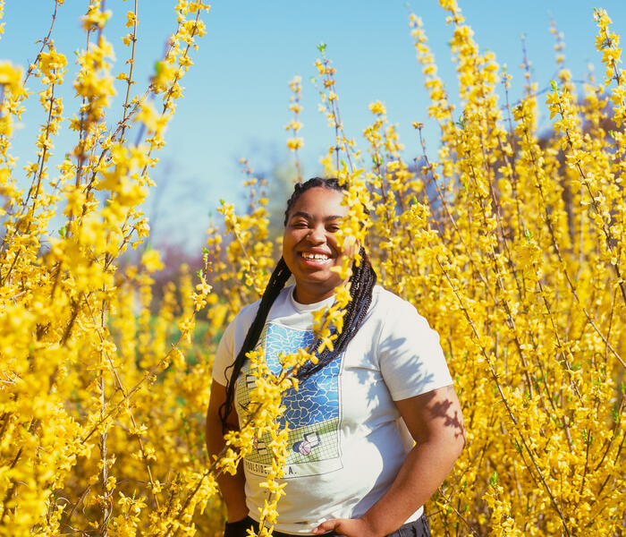 Singer NAE smiling with Joy in a field of yellow flowers surrounding her.