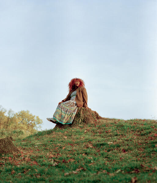 Singer Nan-Ana sitting on a tree stump on top of the hill with a blueish gray sky behind her.