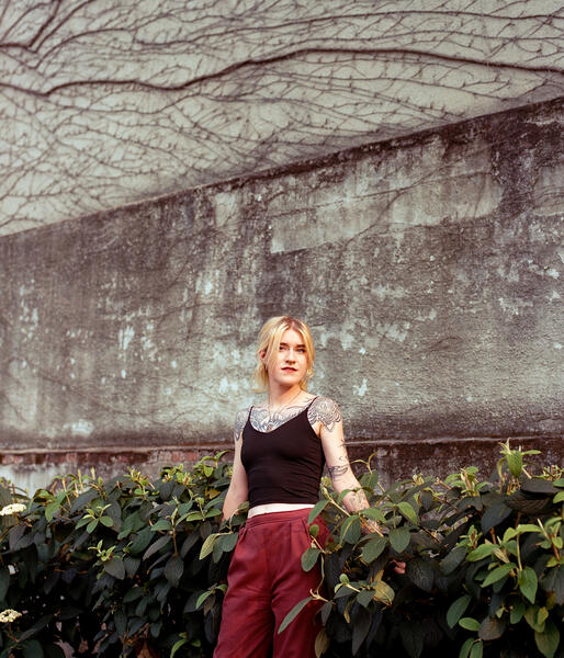 Illustrator Evangeline standing and posing with her arms spread out into the bushes on either side of her. Behind her is a ivy vine filled wall creating vain like forms on the wall.
