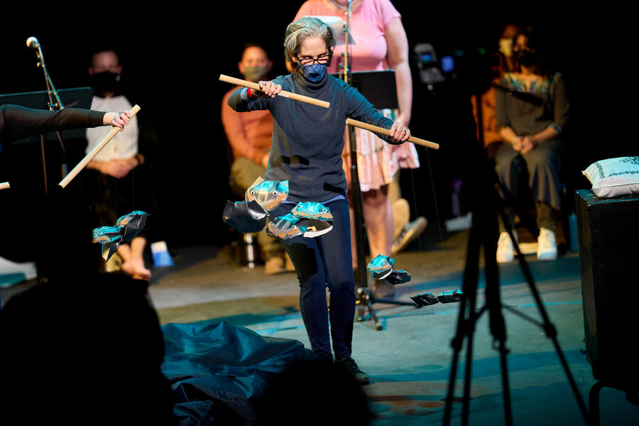 Woman operates paper oyster puppets connected to a rod by string. Three perfomers are seated in the backround, and one performer stands to deliver text for the play reading..
