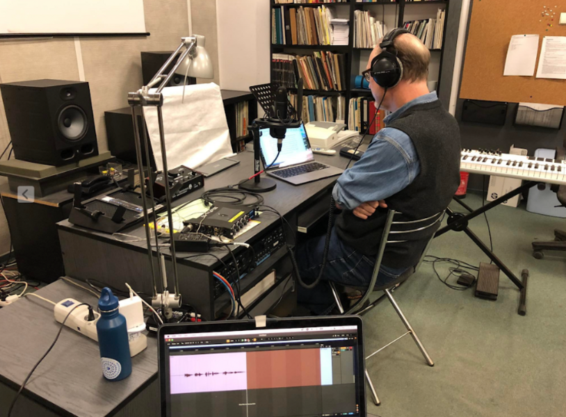 Michael Downs, photographed from the side and behind, seated and recording a podcast. Around are all sorts of audio equipment. He wears headphones.