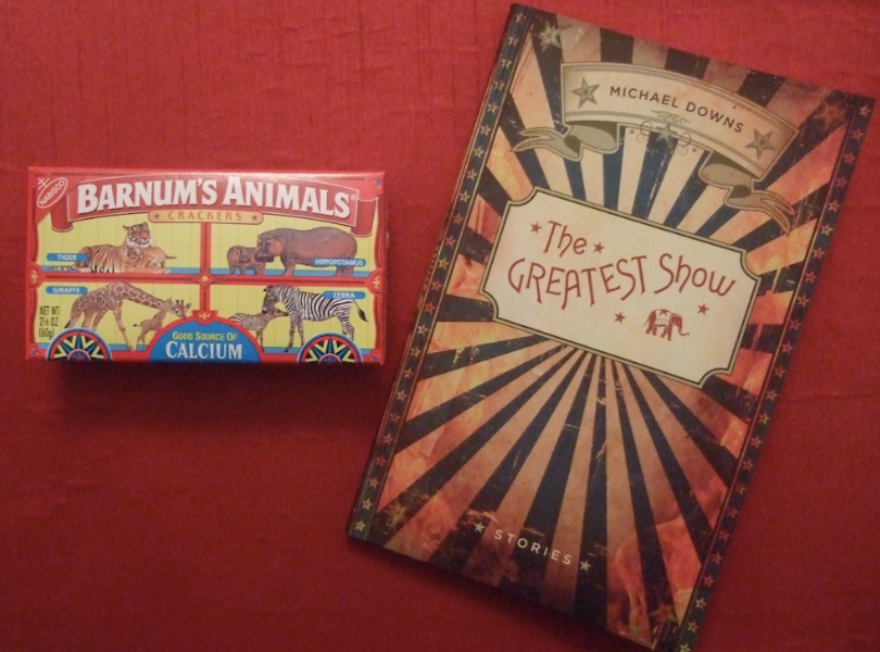 A box of circus animal crackers accompanies a book, The Greatest Show, with a cover that imitates an old circus sandwich board, green and yellow arrows radiating from a placard at the center. The placard holds the book's title and an elephant. Barely visible are flames licking up from the bottom of the image.