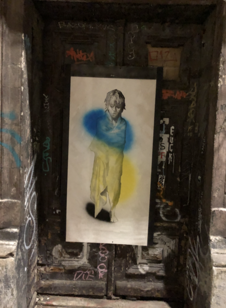A doorway to a tenement house, decorated with graffiti. Pinned to the doorway, on a white background, is the image of a boy, and overlaying that image are the blue and yellow of Ukraine, sprayed with aerosol paint.