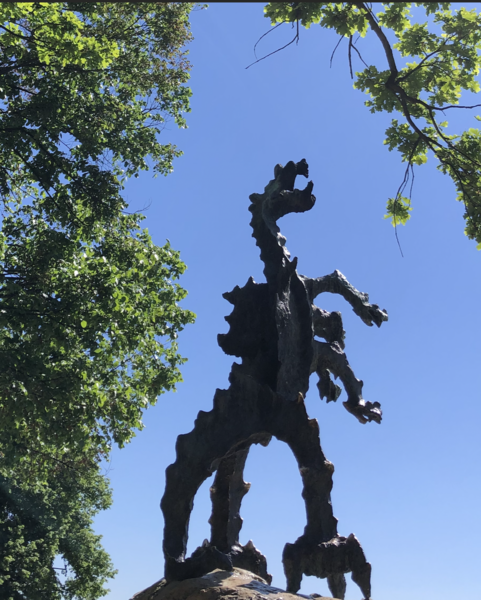 Framed by trees, a metal, jagged sculpture of a dragon rears on its two back legs to roar at a blue sky.