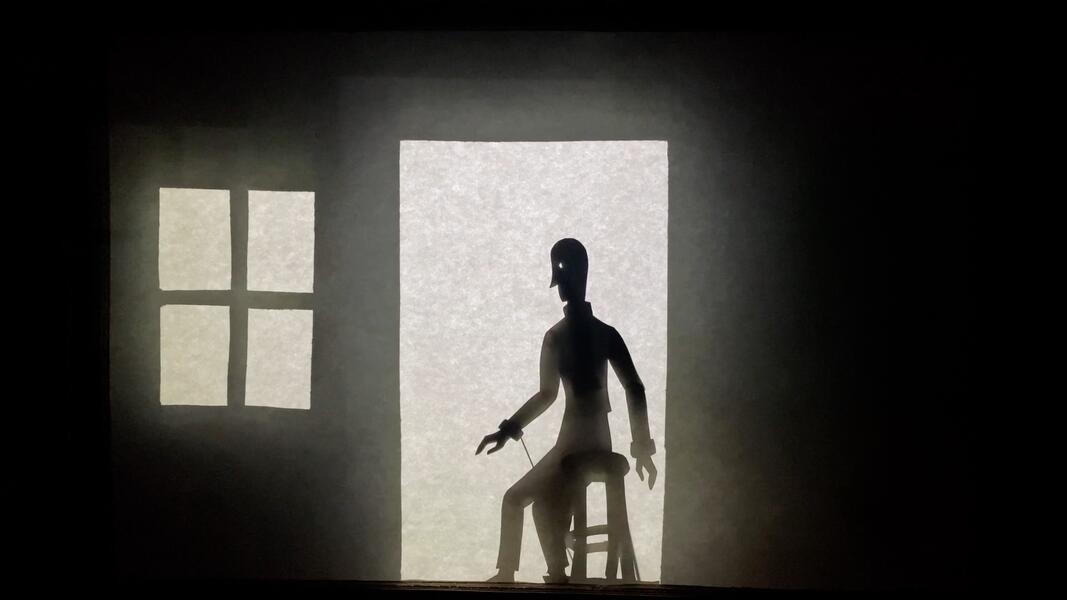 A shadow puppetry setting with a person seated on a stool and a window nearby.