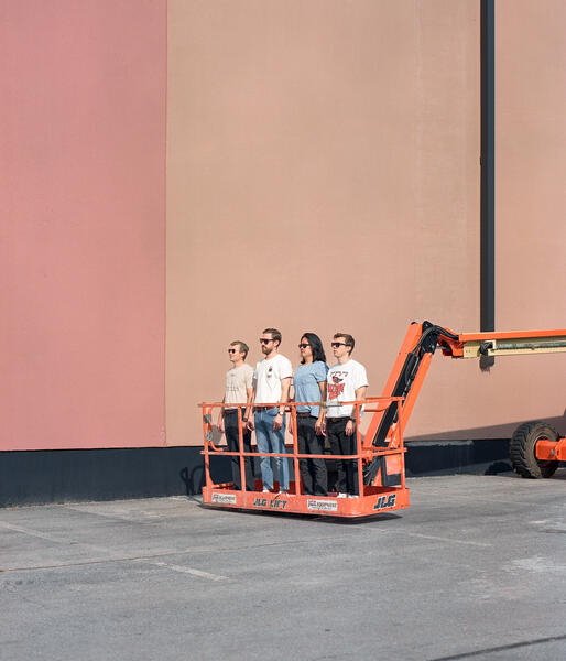 The band Thunder Club stands stiff in a perfect 45 degree angle on the cart of a cherry picker. The Cherry picker is on ground level. The Machine is bright orange infront of a multi tones of red and orange wall