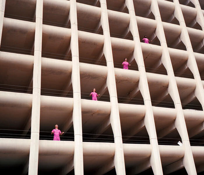 Band Powerwasher standing at the openings of different levels of a parking garage while wearing pink jumpsuits. Photographed from below.