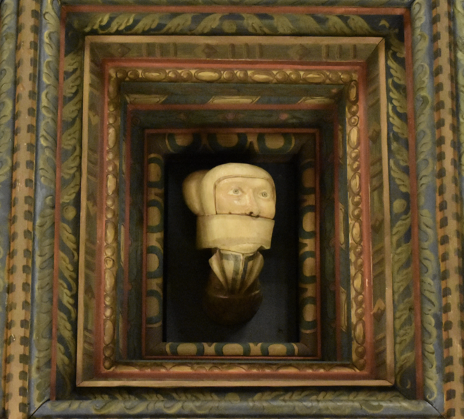 A decorative frame at the center of which is a polished and painted carving of a medieval woman wearing a linen cap and with a white strip of cloth wrapped around her face, covering her mouth.