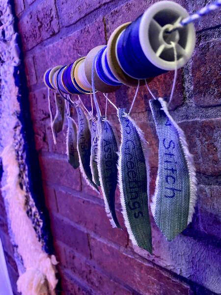 Spools of blue thread hang on a braided blue cord. Each spool has a small piece of denim attached, with numbers and words written on them (e.g. 1 Stitch).