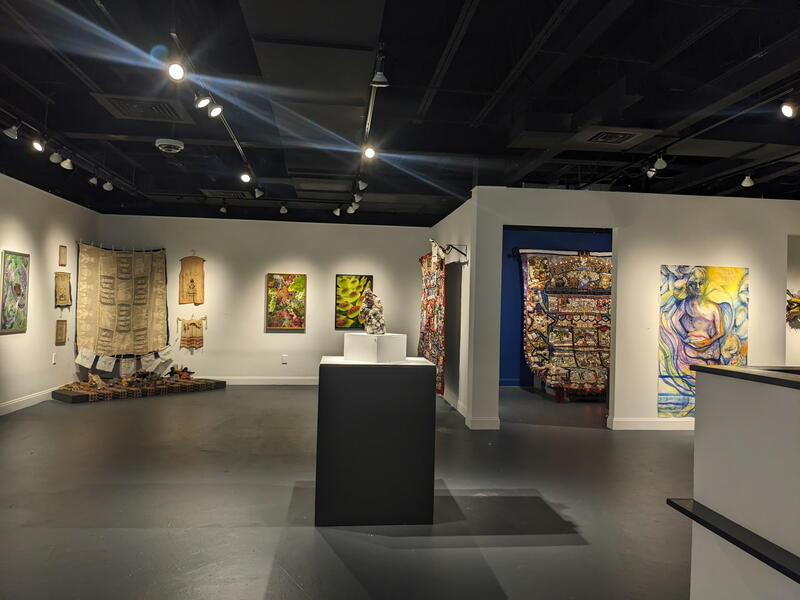 The view just inside the main gallery room. Ceramic on pedestal by Jane Kelly Morais is in memoriam. The exhibit is dedicated to Jane who passed away in April of 2025. Her ceramic is titled Grandma Flew the Coop. Corner installation by Dr Leslie King-Ha