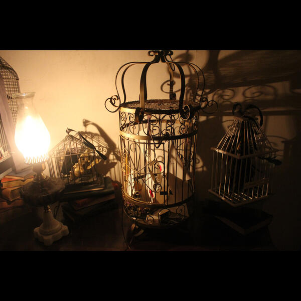 Three bird cages sit atop a table. One is filled with hand written letters hanging from ribbons, and the other two contain assembleges of architectural hardware.