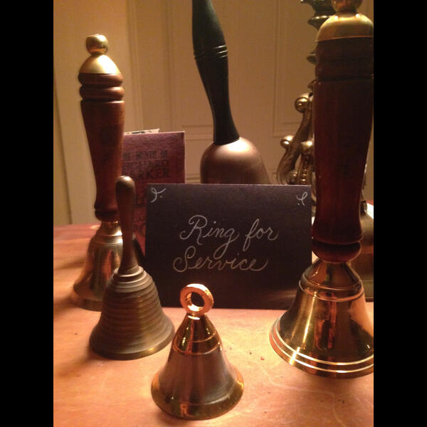 A variety of hand bells surround a hand written sign that reads "Ring for Service" on a bar top.