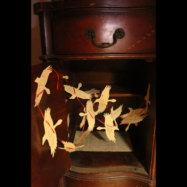 An open cabinet reveals crow silhouettes made of paper and connected by red ribbon inside.