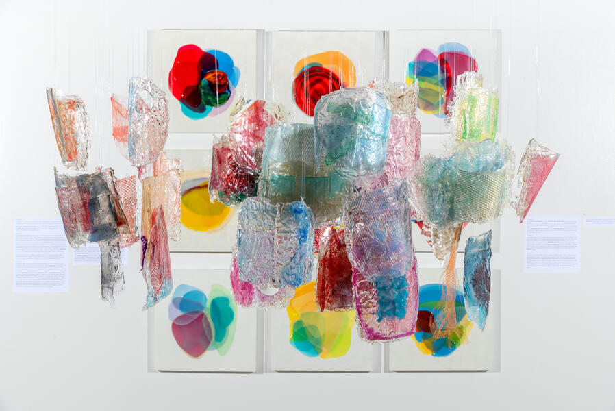 Hanging resin installation in front of wall of colorful paintings by Farida Hughes