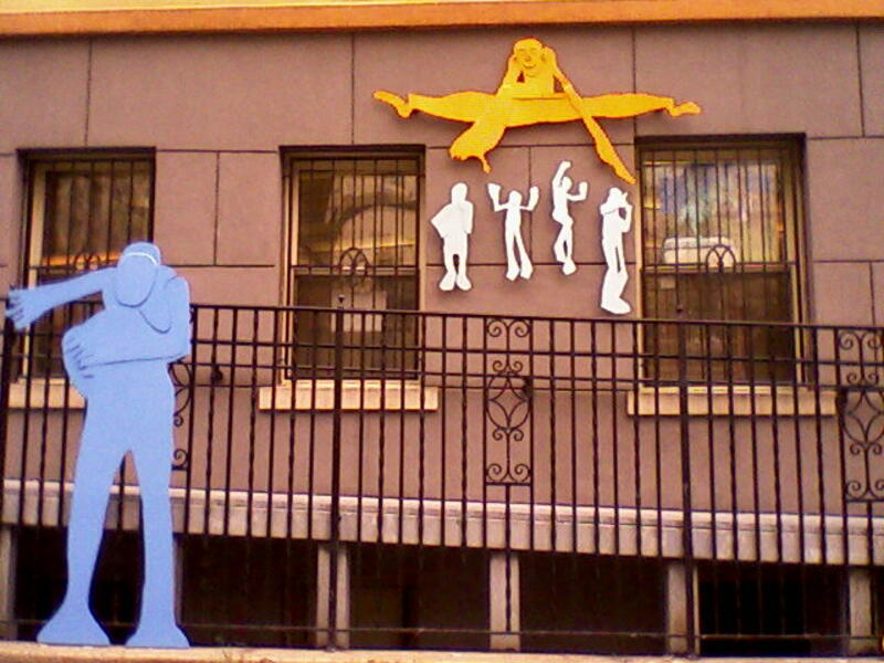 WVSA;School for the Arts in Learning (detail); one flat 7 ft tall figure on the left , then hanging on the wall is a yellow flat metal cut out figure doing a jumping split (about 4 feet across), with 4 small white figures ( flat metal cutouts) underneath.