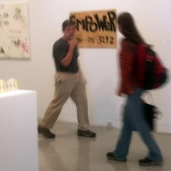 A sign painted on a piece of wood that says EMPOWER with the artist's phone number. It is hanging in the art gallery and people are walking by it.  