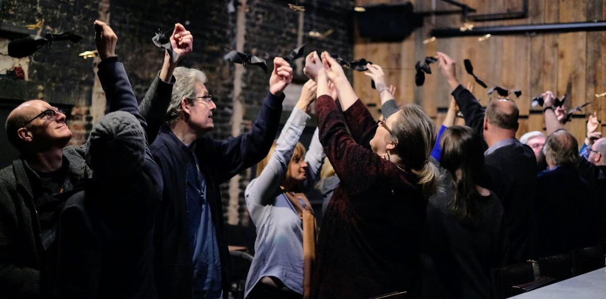 Audience members hang black paper shaped like bats from a suspended string in a bar.