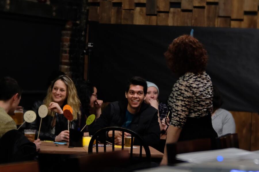 A woman performer talks with audience members who are seated at tables in a bar.