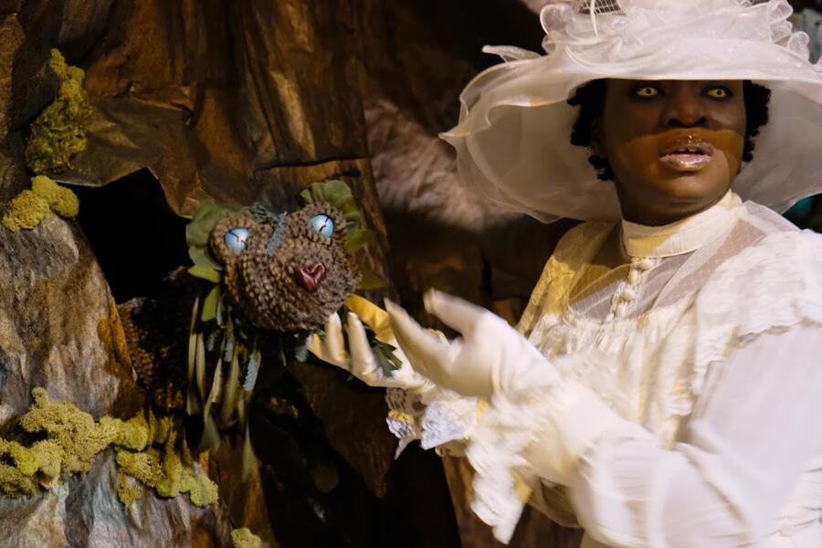 A woman performer with yellow eyes and wearing a fancy white hat and dress touches a puppet that emerges from a cave-like wall. The puppet, representing an otherworldly species, appears to be a cross between a snake and a bear with blue eyes and a leafy mane.
