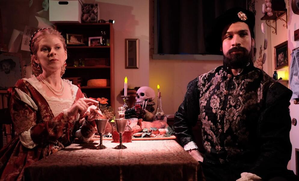 A male and female performer, wearing costumes from the Renaissance period, are seated opposite one another at a table. The table is covered with candles, a banquet, and wine cups. The woman has a ring with a secret compartment open and is pouring something from it into one of the cups of wine.