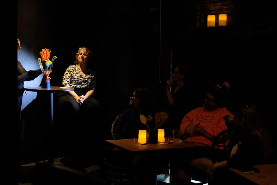 A tabletop puppet, with orange and red hair standing straight up and bulging yellow eyes, representing generalized anxiety, sits atop a bar table. A woman performer seated next to the table looks at the puppet. Audience members seated at bar tables look on.